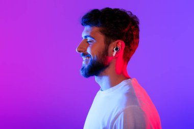 Kulaklıklı genç bir adamın profil portresi çevrimiçi müzik dinliyor, gözleri kapalı poz veriyor mor neon stüdyo arka planında kablosuz kulaklık takıyor, müzik listesinin tadını çıkarıyor.