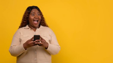 Bin yıllık heyecanlı Afro-Amerikan bayan elinde akıllı telefonuyla boş fotokopi alanına bakıyor ve sarı stüdyo arka planında izole edilmiş olduğunu iddia ediyor.