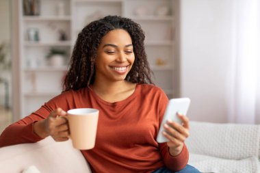 Genç Afrikalı Amerikalı kadın akıllı telefon ve kahveyle koltukta dinleniyor, cep telefonu kullanan siyah bir kadın gülümsüyor ve evde dinlenirken sıcak içkinin tadını çıkarıyor.