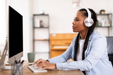 Beyaz kulaklıklı konsantre siyahi kadın özenle bilgisayar başında çalışıyor. Ev öğrenme ortamı içinde online bir derse dalmış.