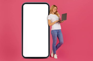 Dijital Freelancer. Laptop bilgisayarında internette sörf yapan sarışın genç bayan, büyük cep telefonunun yanında boş ekran ile dururken, stüdyoda pembe arka planla karşılaşıyor. Web sitesi ya da uygulama için model