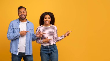 Güzel teklif. Parmakla gösterilen mutlu siyah çift, fotokopi alanını gösteriyor, genç Afrikalı Amerikalı erkek ve kadın reklamınız için boş yer gösteriyor, sarı arka planda duruyor,