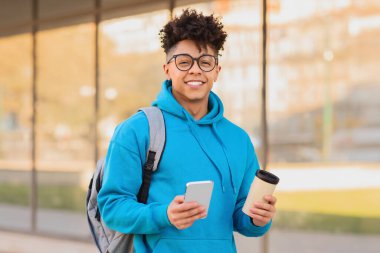 Neşeli Brezilyalı öğrenci cep telefonu ve modern kolej binasının dışında kahvesini yudumluyor, sırt çantasıyla şehirde poz veriyor, kameraya gülümsüyor. Öğrencilerin yaşam tarzı