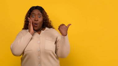 Bin yıllık tombul Afro-Amerikan kadın bej renkli jestler yapıp reklam için boş fotokopi alanı gösteriyor, yüzüne dokunuyor, sarı arka planda izole edilmiş.