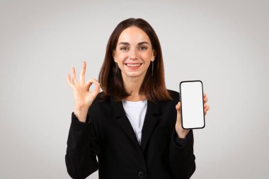 Neşeli Kafkasyalı bin yıllık iş kadını boş bir akıllı telefon ekranı gösteriyor ve onay işareti yapıyor, ya da mobil bir uygulama ya da servisin olumlu bir eleştirisi