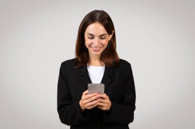 Klasik siyah takım elbiseli bin yıllık beyaz iş kadını, akıllı telefonuyla meşgul, muhtemelen hoş bir mesaj okuyor ya da işle ilgili bilgileri inceliyor.