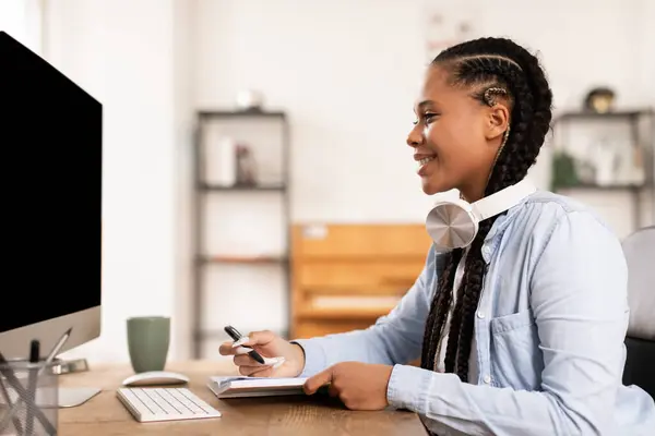 ノートを取るようにヘッドフォンで笑顔に魅了された黒人女性は 自宅で彼女のコンピュータ上でインタラクティブで楽しいEラーニングの経験を示唆しています — ストック写真