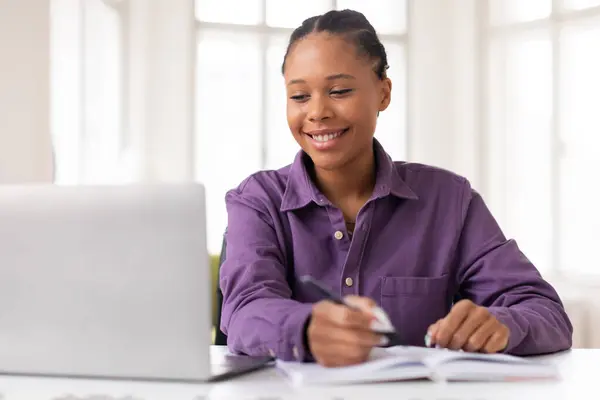 ハッピー アフリカ系アメリカ人 ティーンエイジャー 紫色のシャツの女性は ノートブックに書き込み 自宅で明るい部屋でラップトップを使用しながら学習を楽しんでいます オンラインレッスンをお楽しみください — ストック写真