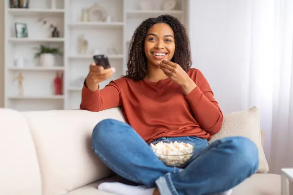 Happy black woman enjoying movie night at home, cheerful african american female holding remote and eating popcorn while sitting cross-legged on comfortable sofa in living room interior, copy space