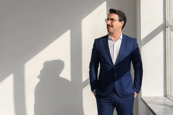 Smiling middle aged european businessman in a sharp blue suit looking away thoughtfully with strong shadows on the wall in a bright, sunlit room, suggesting optimism and vision