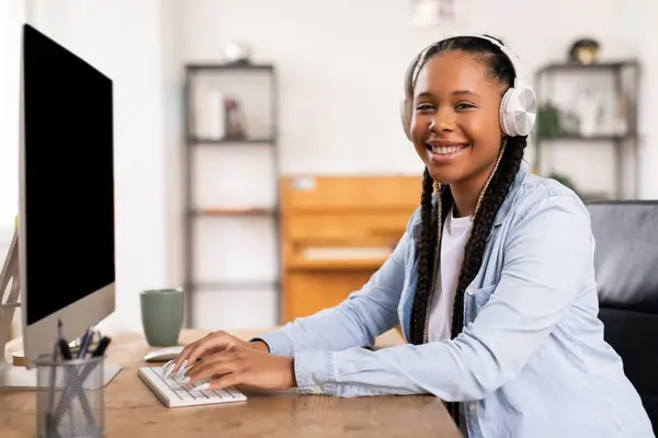 快乐的黑人女学生 带着耳机笑着打字 积极参加家里的在线教育活动 — 图库照片