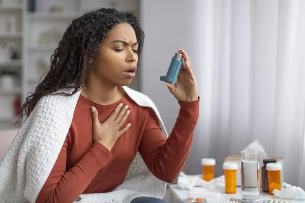 Young black woman experiencing an asthma attack at home, using an inhaler for relief, sick african american female clutching her chest in distress, sitting at table with medication, free space