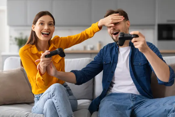 Excited woman playfully covers mans eyes, both with gaming controllers, laughing and enjoying playful gaming session on their comfortable couch at home