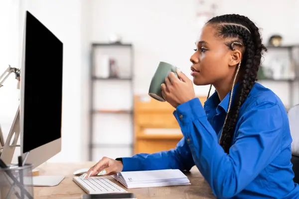 专心致志的黑人女生穿着衬衫 一边喝着咖啡 一边在她整洁整齐的电脑桌上工作 桌上还配备了黑色监视器 — 图库照片