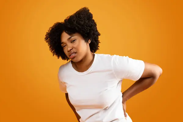 アフリカ系アメリカ人ミレニアル世代の女性が背中に触れた白いTシャツを着て 期間中に筋肉が痛み オレンジ色のスタジオの背景に苦しんでいます — ストック写真