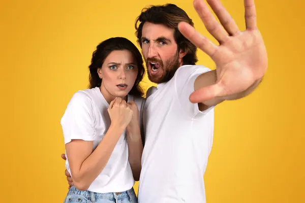 A frightened european woman and a protective man with a beard express fear and defense, with the man reaching out as if to stop something, against a yellow backdrop, studio