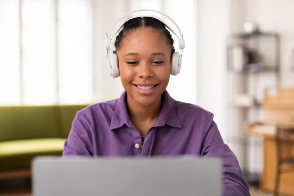 笑顔黒十代の女性がオンライン学習に従事し ホワイトヘッドフォンを着用し ラップトップを使用して 自宅の設定でカジュアルな紫色のシャツを着て — ストック写真