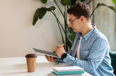 Kafkasyalı, kıvırcık saçlı ve gözlüklü bir öğrenci dijital tablet ve stil kullanarak bir proje üzerinde çalışıyor, bitki dolu bir odada kahve fincanı masanın üzerinde.