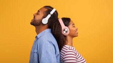 Kablosuz kulaklık takan, parlak sarı arka planda sırt sırta duran, gülümseyen, sakin, Afro-Amerikan erkek ve kadın favori müziği dinleyen rahat siyah bir çift.