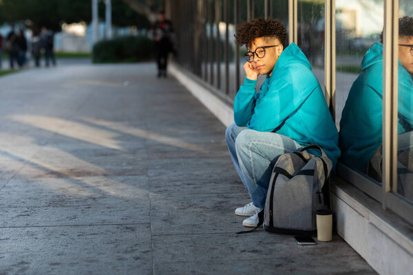 Несчастный чернокожий студент, сидящий с рюкзаком на открытом воздухе в городской местности ночью, исключенный из колледжа, имеющий проблемы с арендой недвижимости, чувствующий себя одиноким и подавленным. Пустое пространство