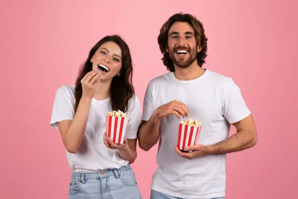 快乐快乐的欧洲女人和男人穿着白色的T恤衫 笑着吃着红白相间的爆米花 在粉红的背景下一起享受着快乐的时光 工作室 — 图库照片