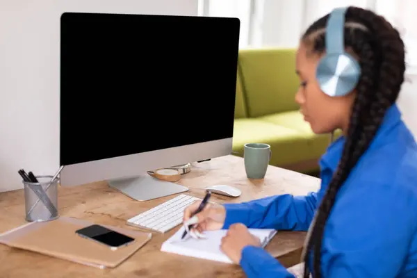 在井井有条的家庭学习环境中 全神贯注的戴着耳机的黑人少女 并在网上电脑课程上做笔记 — 图库照片