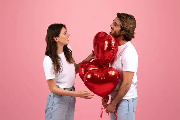 热情的高加索年轻夫妇 带着柔和的粉红画室背景 带着浪漫和幸福的色彩 嬉笑地握着并看着闪闪发亮的红色心形气球 — 图库照片