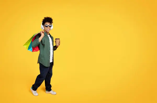 时尚时尚的年轻非洲裔美国人 在购物时戴着太阳镜和无线耳机听音乐 背着五颜六色的纸袋和外卖咖啡 黄色背景和复制空间 — 图库照片