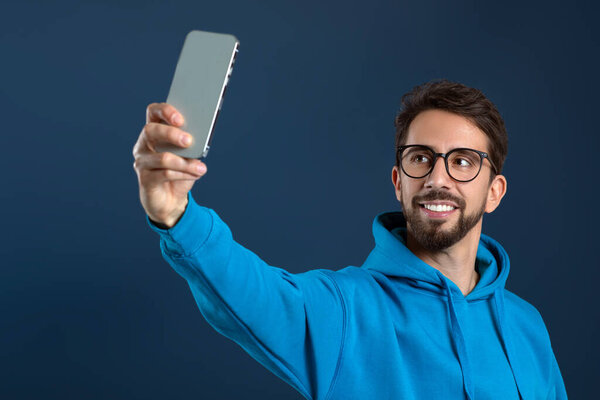 Handsome young man in eyeglasses taking selfie on smartphone, happy millennial guy smiling at phone camera, standing against dark blue studio background, creating content for social media