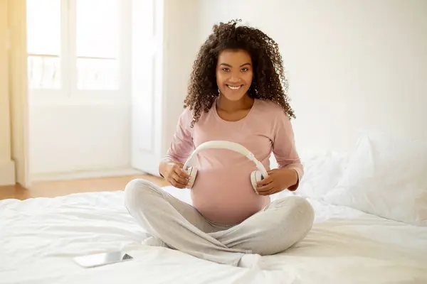 Happy black pregnant woman holding headphones near belly, playing melody to baby in womb, african american expectant mother listening classical music during pregnancy, sitting on bed at home