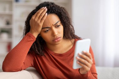Endişeli genç siyahi kadın akıllı telefon ekranına bakıyor ve kafaya dokunuyor, üzgün Afrikalı Amerikalı kadın kanepede oturuyor sıkıntılı bir ifadeyle, stresle ya da telefonda kötü haberler alıyor.