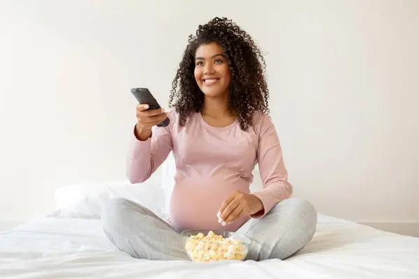 Pregnant black woman in pink shirt enjoying popcorn and holding remote controller, african american mother-to-be watching tv while sitting comfortably on bed, relaxing in bedroom at home