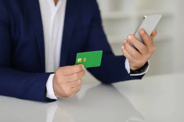Close-up of middle aged european businessmans hands holding a green credit card and a smartphone, likely managing online banking or conducting a mobile transaction, cropped