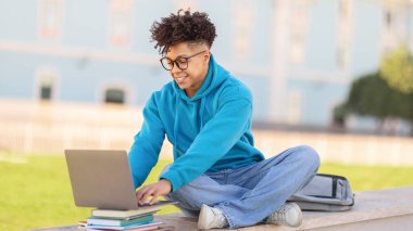 Çevrimiçi Çalışma. Bilgisayarda yazı yazan ve internette gezinen gülümseyen zenci öğrenci. Üniversite kampüs parkında dışarıda oturarak öğreniyor. Teknoloji ve üniversite eğitimi, E-öğrenme kavramı. Panorama