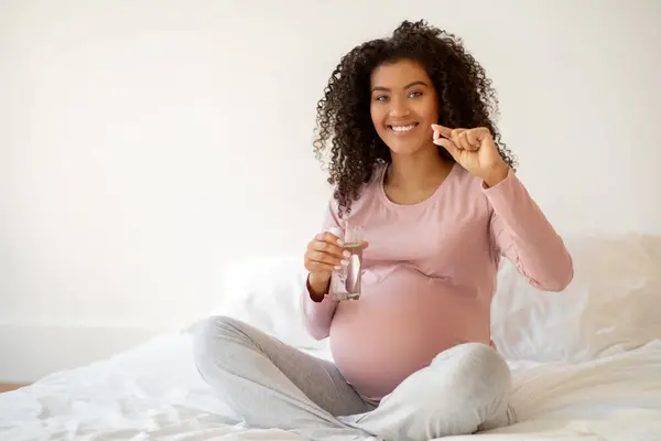 Smiling black pregnant woman showing vitamin pill and holding glass of water, african american expectant mother promoting health and prenatal care while sitting on bed in cozy bedroom at home