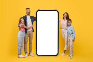 Dört kişilik mutlu bir aile, boş ekranlı büyük akıllı telefonun yanında duruyor, gülümseyen genç ebeveynler ve onların çocukları mobil reklam uygulaması, sarı stüdyoda birlikte poz veriyorlar, kolaj,