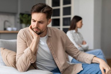 Endişeli adam koltukta oturuyor, elleri yanaklarında, derin düşüncelere dalmış, kadın ise arkasını ona dönmüş, ilişki zorluklarını tasvir ediyor.