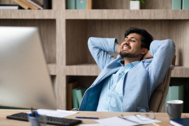 Rahat Hintli işadamı arkasını yaslıyor ve ellerini başının arkasına koyuyor, modern ofis iç bilgisayarının yanında otururken işe ara veriyor.