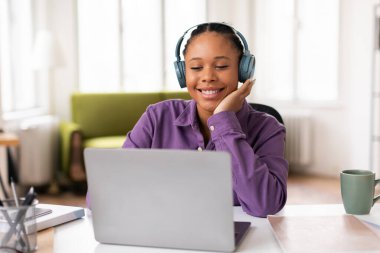 Kulaklık takan, mor tişörtlü neşeli siyah kız, online eğitime dizüstü bilgisayarla başlıyor, yeşil kanepeli iyi ışıklandırılmış bir odada.