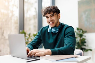 Boynunda şık kulaklıkları olan neşeli genç adam çalışırken ya da bilgisayar başında çalışırken gülümsüyor.