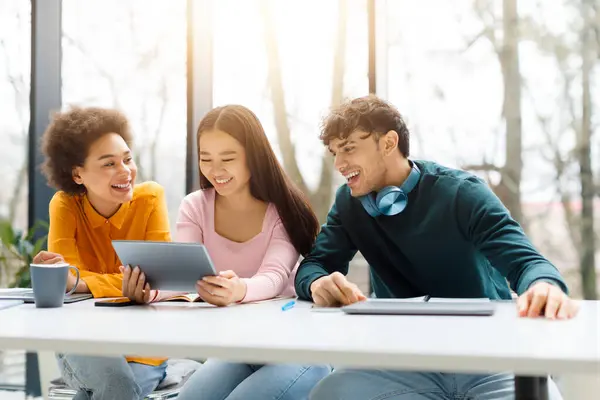 Joyful young diverse students sharing tablet and enjoying coffee while studying together in sunlit room filled with laughter and productivity, free space