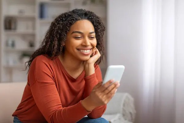 Smiling Black Woman Reading Message On Smartphone While Sitting On Couch At Home, Happy Young African American Female Using Mobile Phone For Texting With Friends Or Online Shopping, Copy Space