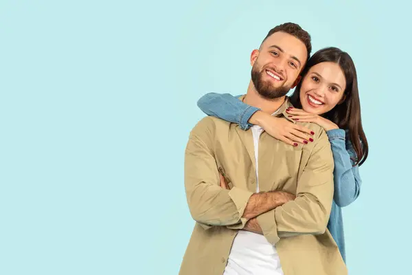 Happy European Young Couple Hugging While Posing Over Blue Studio Backdrop, Standing Near Copy Space For Advertisement Text, Smiling To Camera. Portrait Of Wife Hugging Husband From Back