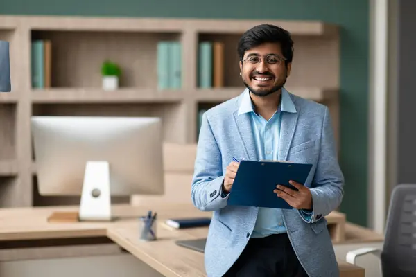 Happy Indian entrepreneur businessman proudly holds clipboard with signed document in office, smiling to camera with confidence, symbolizing successful deal. Business offers, career