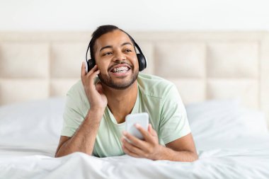 Yakışıklı, sakallı, Afro-Amerikalı adam rahat yatak odasında dinlenirken, telefon ve kablosuz kulaklık kullanarak fotokopi odasına bakıyor ve gülümsüyor.