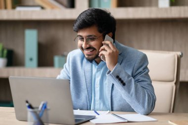 Orta Doğulu iş adamı telefonla iletişim kuruyor ve ofisteki masasında dizüstü bilgisayar ve akıllı telefonla görevleri yönetiyor. Modern çalışma ve iletişim