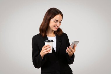 Milenyum Kafkasyalı, siyah takım elbiseli bir iş kadını elinde tek kullanımlık bir kahve fincanı tutarken mutlu bir şekilde telefonunu kontrol ediyor, verimliliği ve bağlantıyı özetliyor.