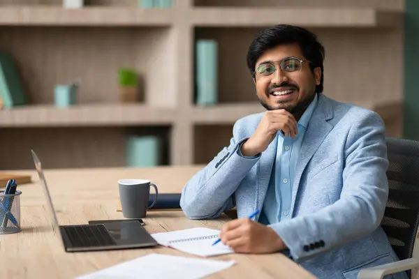 Indian executive businessman sitting with happy smile while taking notes, comfortably working at modern laptop PC at office indoor, smiling to camera with confidence. Successful business career