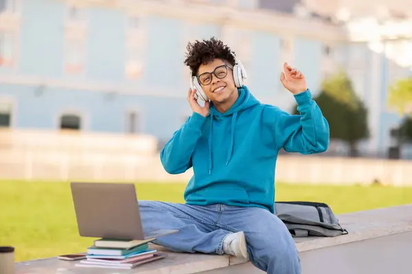 Cheerful African American college student guy wearing wireless headset finds joy in music, sitting with laptop during break at university campus park, learning and relaxing outdoors