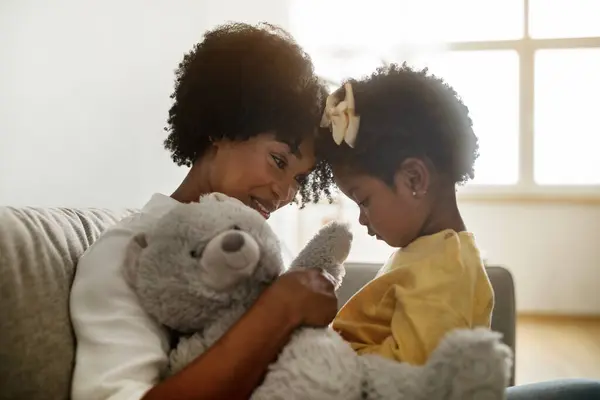 Family time concept. Cute curly little kid african american girl playing with her mother, happy black toddler child and mom sitting on couch, holding teddy bear, home interior, closeup